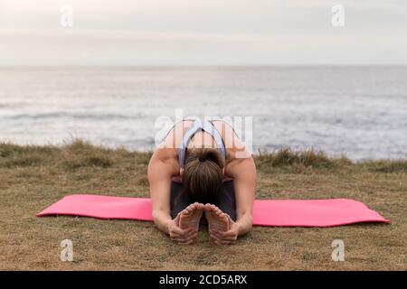 Woman in sportswear stretching outdoors on a pink mat with the sea in the background. Stock Photo