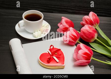 Holiday concept. Bouquet of pink tulips, a cup of coffee, red heart-shaped cookies with a note, empty magazine on a black wooden background. Stock Photo