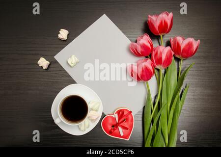 Holiday concept. Bouquet of pink tulips, a cup of coffee, red heart-shaped cookies with a note marshmallow and sheet of paper on a black wooden backgr Stock Photo