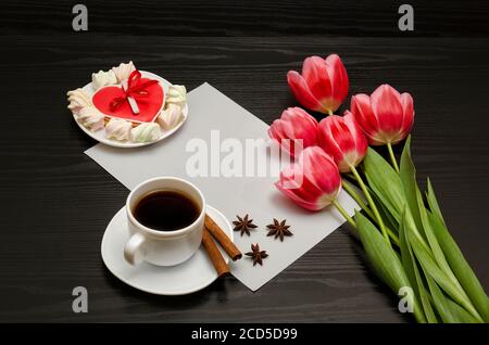 Holiday concept. Bouquet of pink tulips, a cup of coffee, red heart-shaped cookies with a note, cinnamon, star anise and sheet of paper on a black woo Stock Photo