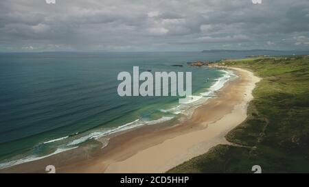 Aerial view sandy beach: ocean foamy waves drops to coastline in White Beach, Antrim County. Seascape of picturesque nature of Northern Ireland. Dramatic summer scenery in day Stock Photo