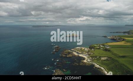 Cliff aerial landscape: rocky ocean shore. Irish plant distillery against backdrop of nature: meadows and cumulus clouds on summer day. Industrial scenery of bird's-eye view Stock Photo