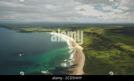 Aerial view sandy coastline, green grass meadows. Waves crashing out of shore and come back to ocean. Atlantic bay serene scenery in dusk summer daytime cinematic shot Stock Photo