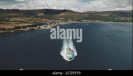 Scotland ocean, passenger ferry aerial view in coastal water of Firth-of-Clyde Gulf. Ship crossing from Brodick terminal to Scottish mainland. Cityscape at green valleys. Cinematic shot Stock Photo