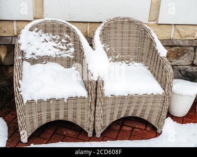 Two wicker chairs and a wicker pot covered with snow stand on a snow-covered tile near a stone wall Stock Photo