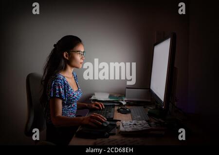 Working late at night on computer. Asian woman at office desk staring at desktop screen wearing blue light eyewear glasses to protect from eye strain Stock Photo