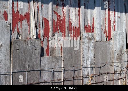 Exterior wood siding on a rustic late 19th Century wooden barn with weathered and worn red peeling paint and rusty metal fencing Stock Photo