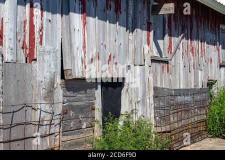 Exterior wood siding on a rustic late 19th Century wooden barn with weathered and worn red peeling paint and rusty metal fencing Stock Photo