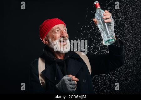 Smiling bum happily look at bottle of alcohol. Homeless male dressed in red hat and black coat stand in the rain isolated over black background Stock Photo