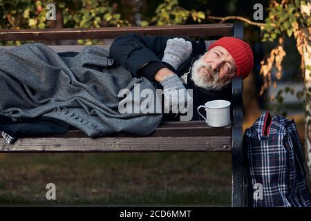 Roofless male lying on street bench asking for money, for any help. Lagguage near bench. Desperate and lonely homeless man without shelter Stock Photo