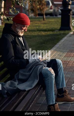 Mature man without shelter sitting in street clothes on bench, without food and money. Drooping man lowered his head down to cup for collecting money. Stock Photo