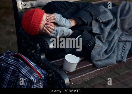 Penniless senior man lying on park bench, trembling from the cold, closed face with hands. Cup for collecting money, coins next to him. Stock Photo