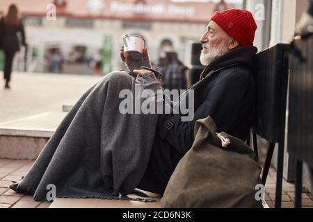Beggar male sit with jar for money, wearing warm old clothes. Vagrant with grey beard, red hat and grey gloves freezing in the street Stock Photo