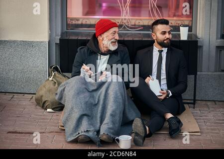 Businessman and beggar male sit eating on street and speaking. Man un tuxedo, poor male in old clothes Stock Photo