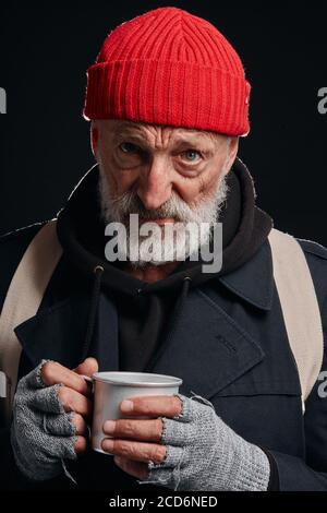 Old bearded vagabond holding mug of hot drink, trying to warm up. Dressed in black coat and red hat, white backpack. Isolated black studio background Stock Photo