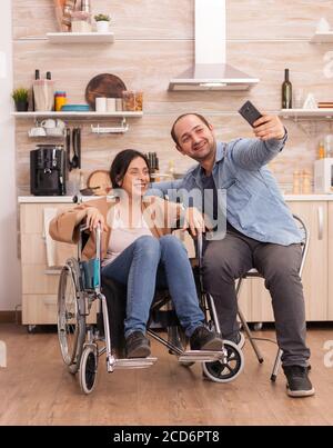Positive woman in wheelchair and husband taking a selfie in kitchen using smartphone. Disabled paralyzed handicapped woman with walking disability integrating after an accident. Stock Photo