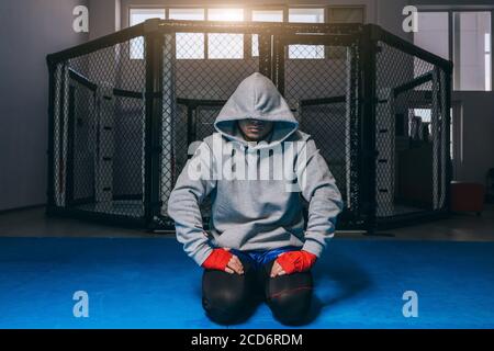 Boxer man in sportswear with hood on head posing on knees in a dark studio background with a boxing ring behind Stock Photo