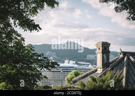 Glover Garden, Nagasaki, Kyushu, Japan - Chimney and roof of the old Japanese house and a huge cruise ship in front of the Mountain Inasa. Stock Photo