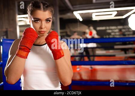 young caucasian MMA girl in sports uniform ready to fight in boxing ring, red bandages on her fists, combat readiness of muscular sportive female Stock Photo