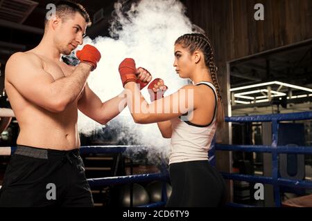 young caucasian man and woman fight with each other in ring, wearing sportive wear, red protective bandages or gloves, isolated over smoky space Stock Photo