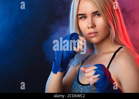 Blonde female athlete is exercising in defense and attacks on dark smoky background. Woman in tank-top and boxing bandages is having cardio boxing wor Stock Photo
