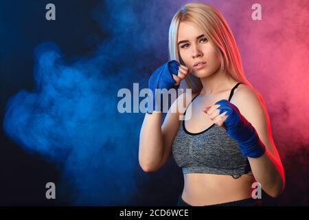 Blonde female athlete is exercising in defense and attacks on dark smoky background. Woman in tank-top and boxing bandages is having cardio boxing wor Stock Photo