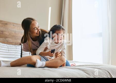 Young mother with her five years old little daughter dressed in casual outfit are relaxing and playing in the bed at the weekend together, lazy mornin Stock Photo