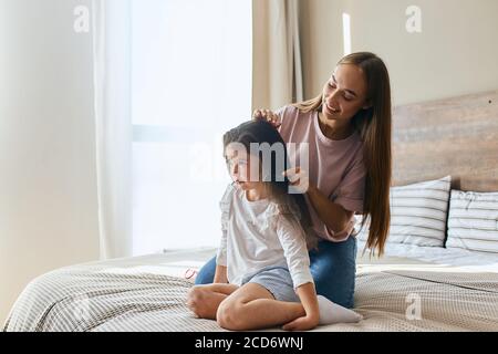 Pretty charming girl with long hair and broad smile holding small hairbrush and combing brunette hair of little daughter sitting on bed, morning time Stock Photo