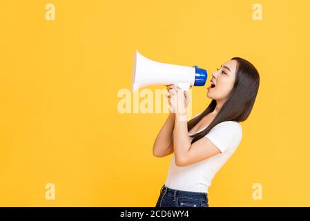 Side view portrait of young pretty Asian woman holding megaphone while shouting announcement in isolated studio yellow background Stock Photo