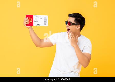 Fun portrait of surprised young Asian man wearing 3D glasses holding movie ticket in isolated studio yellow background Stock Photo