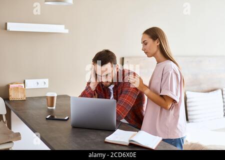 Upset frustrated male in optical glasses, casually dressed, sits at black wooden table stares at computer with dissatisfied feeling, terrified wife st Stock Photo
