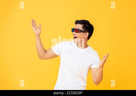 Fun Portrait of excited young Asian man wearing 3D cinema glasses with both hands raised in isolated studio yellow background Stock Photo