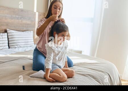 Young mother casually dressed sitting on knees on big bed, combing curly hairs of little brunette daughter, preparing for kindergarten, family portrai Stock Photo