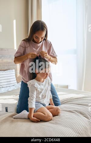Pretty mother and little daughter getting ready to go to kindergarten, woman combing long hair of brunette kid, putting scrunchy, in bed room, posing Stock Photo