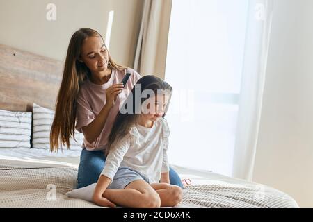 Beautiful charming girl in home clothes, sitting on knees on big bed together with mom, looking aside with bright eyes and calm face, posing against b Stock Photo