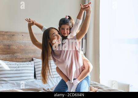 Charming cheerful four years old girl sitting on back of playful mother, looking at camera, spreading heads sideways, happy to be with mom, warm and c Stock Photo