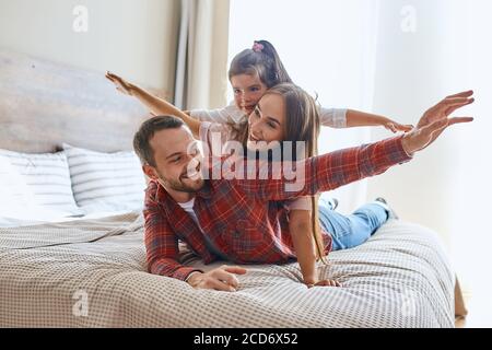 Joyful playful family resting on big wide bed in brightly lighted hotel room, playing with cheerful expression, having fun, enjoying holidays, stretch Stock Photo