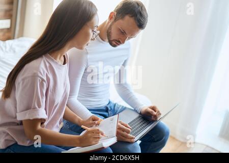Attractive charming lady in round glasses, making notes, holding pencil, turning head at young unshaved boyfriend, planning holidays, indoor shot, clo Stock Photo