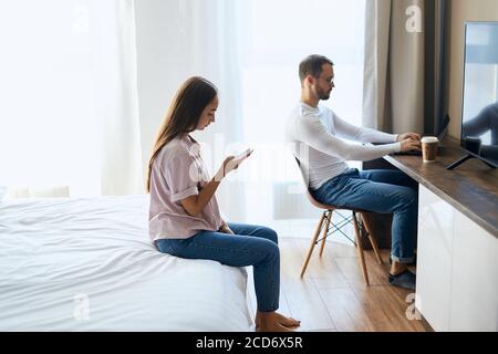 Tall young boyfriend sitting in front of laptop computer, working hard with calm face, pretty girlfriend chatting with friends using phone behind seri Stock Photo