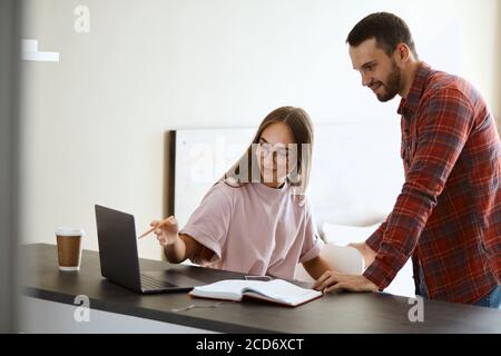 charming playful girlfriend points at screen of laptop computer, showing funny photo, sitting at black wooden table, friendship concept Stock Photo