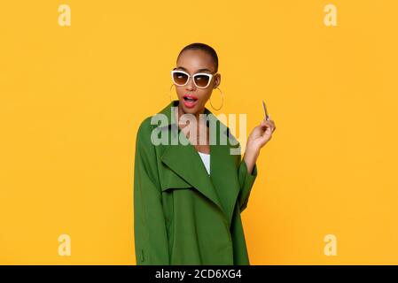 Portrait of happy astonished young fashionable African American woman holding credit card in isolated studio yellow background Stock Photo