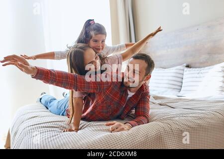 Happy loving family spending time together, father, mother and daughter playing cheerfully on big bed in hotel room, stretching hands, having fun, smi Stock Photo