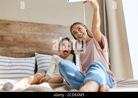 Happy loving family. mother and child girl playing, having fun, taking photo, showing tongue, smiling brightly, morning time in bedroom, shot from bel Stock Photo