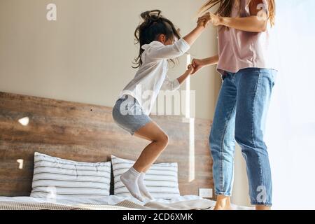 Portrait of little joyful six years old girl happily jumping on bed together with sister in brightly lighted bedroom, shot from below, family joy conc Stock Photo