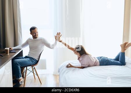 Young bearded man in optical glasses, working at laptop computer in bedroom, turning towards joyful girl, stretching hands, clapping with palms, showi Stock Photo