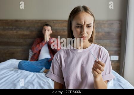 Close up of frustrated woman with pregnancy test in hand, knitting her brows with shocked expression, demonstrating fear and sadness, background of ma Stock Photo