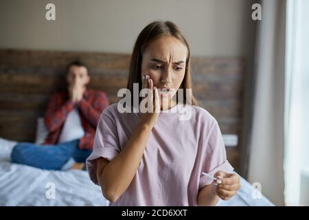 Frustrated sad female standing in bedroom, staring at pregnancy test with opened mouth, raising hand towards cheek with surprised expression, demonstr Stock Photo