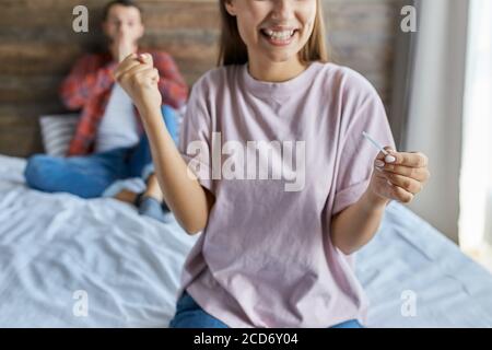 Charming young lady happy to find out about pregnancy, looking at test with toothy smile, makes yes gesture, surprised man sitting behind happy wife, Stock Photo