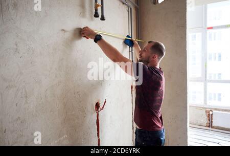 Side view of serious man using measuring tape in room with underfloor heating pipes. Designer in shirt taking measurements while working on renovation of apartment.