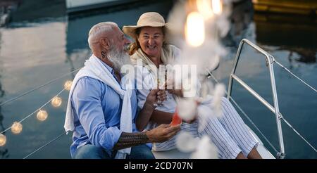 Senior couple toasting champagne and eating fruits on sailboat vacation - Happy elderly people having fun celebrating wedding anniversary on boat trip Stock Photo